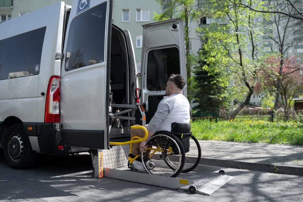 A person in a wheelchair is being assisted onto a accessible van using a lift, surrounded by greenery and residential buildings.