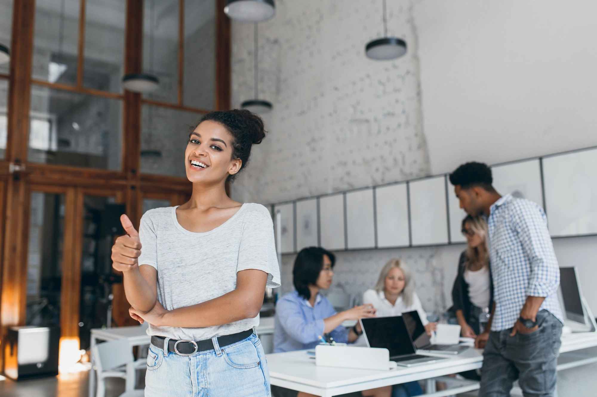 A woman gives a thumbs-up in a modern office environment, with colleagues collaborating at a table in the background